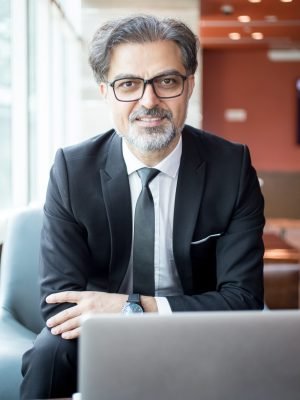 Closeup portrait of smiling middle-aged handsome business man looking at camera and sitting at table with laptop computer in lobby. Front view.