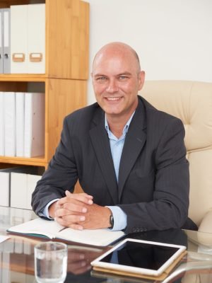 Portrait of balded psychologist specialist sitting at the table with notepad and touchpad and smiling at his cabinet